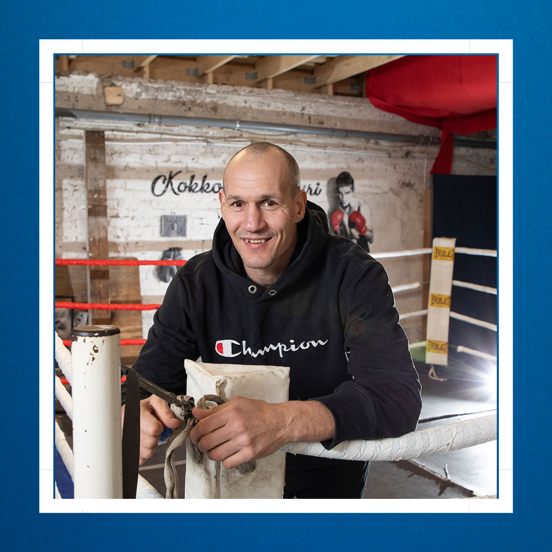 Nyrkkeilijä/valmentaja Amin Asikainen nojaa nyrkkeilykehän köysiin ja hymyilee.